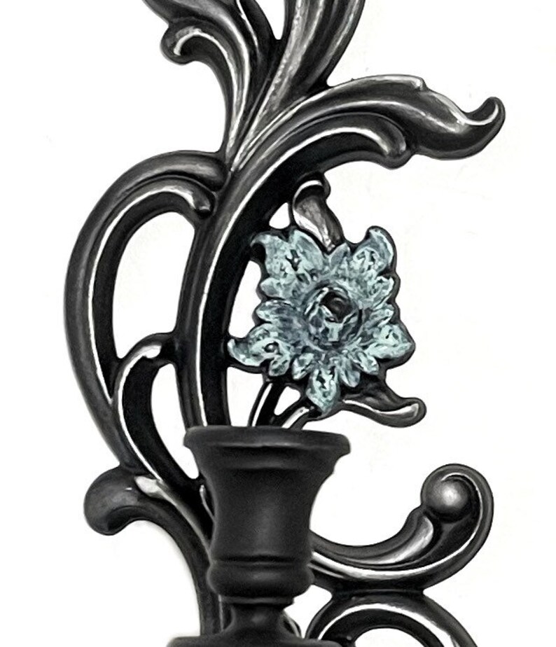 May include: A black metal wall sconce with a single candle holder. The sconce features a decorative floral design with a blue and white flower.