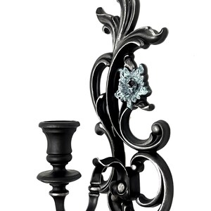 May include: A black metal wall-mounted candle holder with a decorative floral design and a single candle cup. The floral design features a blue and white flower.