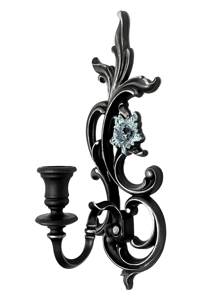 May include: A black metal wall-mounted candle holder with a decorative floral design and a single candle cup. The floral design features a blue and white flower.