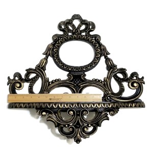 May include: A gold and black ornate wall shelf with a decorative oval frame at the top. The shelf is approximately 11 inches wide.