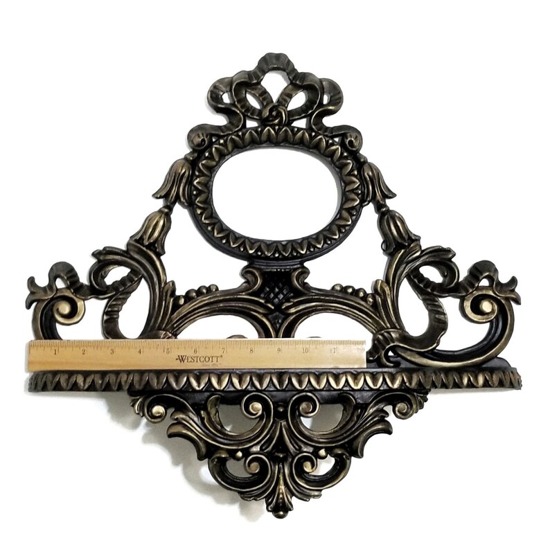 May include: A gold and black ornate wall shelf with a decorative oval frame at the top. The shelf is approximately 11 inches wide.