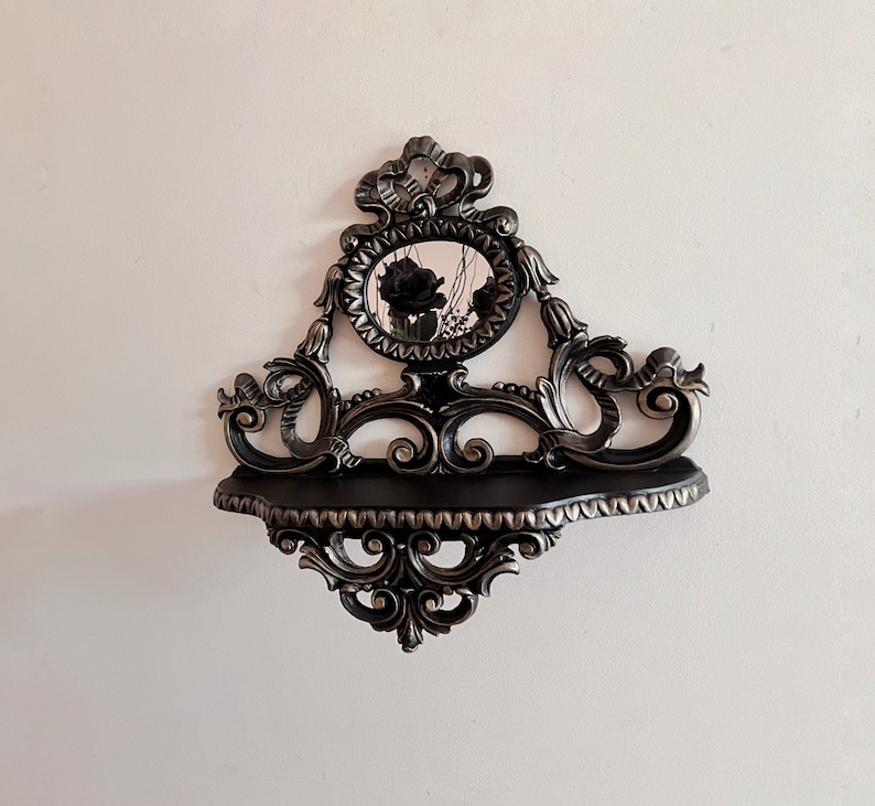 May include: A black and silver ornate shelf with a round mirror in the center. The mirror has a black frame and a floral design. The shelf has a decorative design with scrolls and swirls.