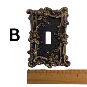 May include: A black light switch plate with a gold ornate frame featuring a floral design with roses. The plate is approximately 3 inches wide.