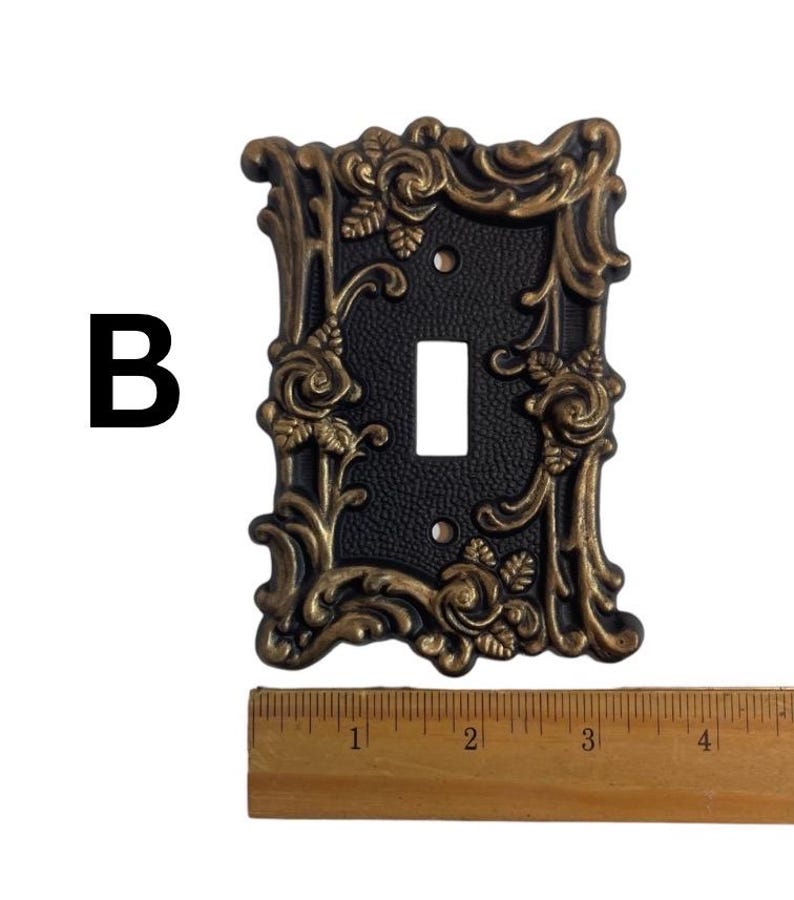 May include: A black light switch plate with a gold ornate frame featuring a floral design with roses. The plate is approximately 3 inches wide.