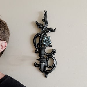 May include: Black metal wall sconce with a floral design and a single candle holder. The sconce has a distressed finish and a blue flower accent.