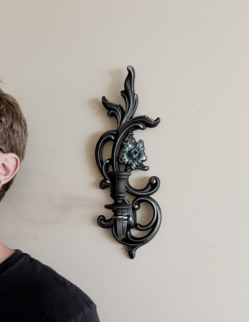 May include: Black metal wall sconce with a floral design and a single candle holder. The sconce has a distressed finish and a blue flower accent.