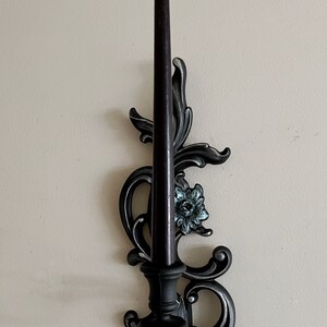 May include: Black metal candle holder with an ornate design featuring a floral motif. The holder has a single candle slot and a black candle is included.