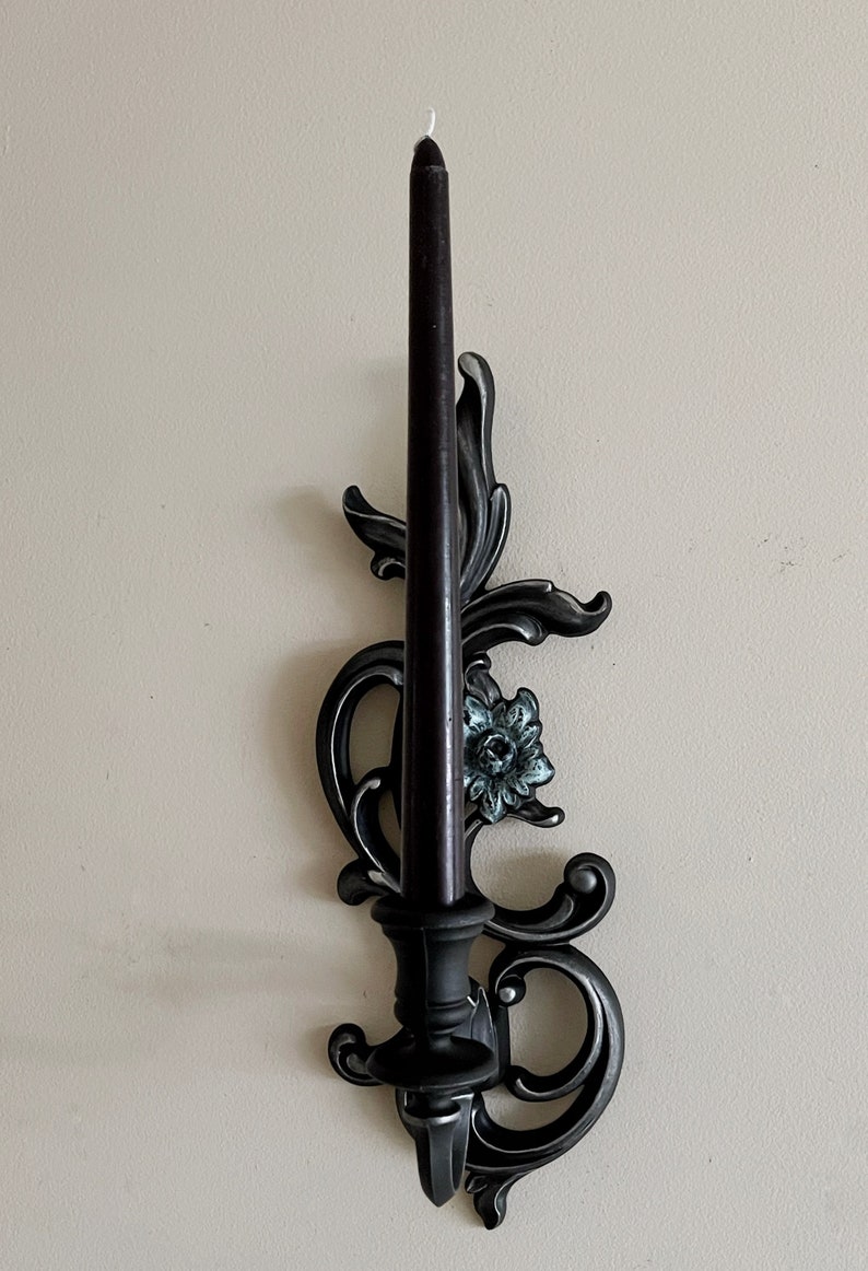 May include: Black metal candle holder with an ornate design featuring a floral motif. The holder has a single candle slot and a black candle is included.