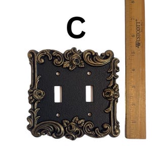 May include: A black double light switch plate with a gold ornate frame featuring floral designs. The plate is approximately 4 inches wide and 5 inches tall.