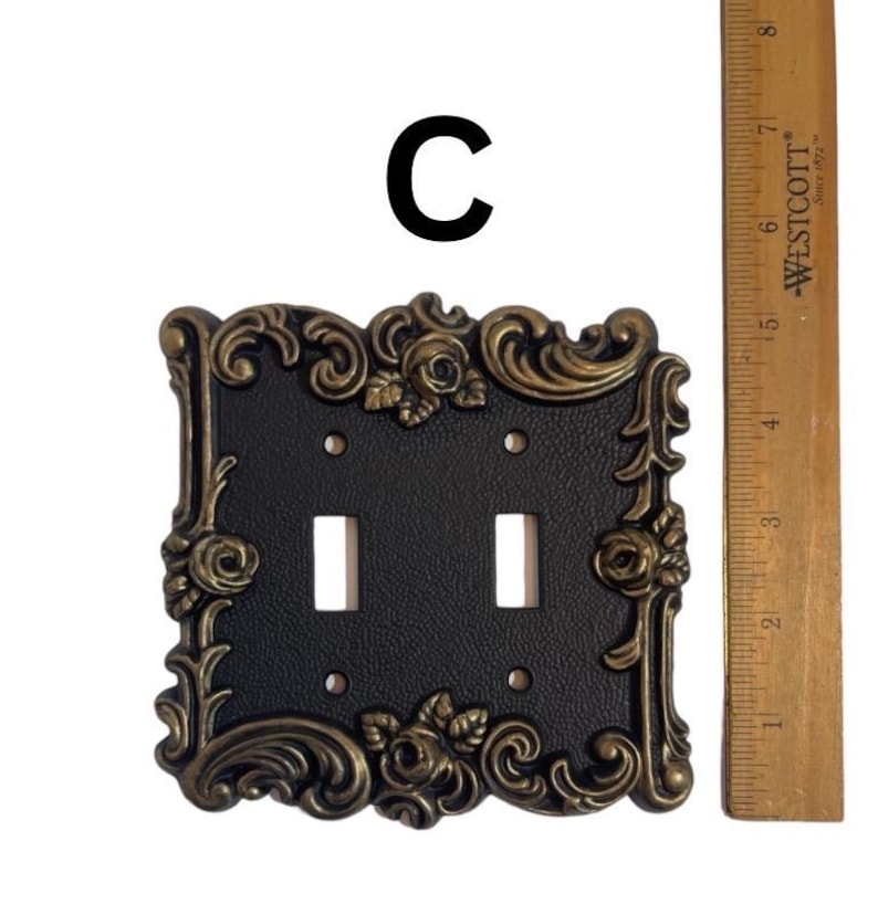 May include: A black double light switch plate with a gold ornate frame featuring floral designs. The plate is approximately 4 inches wide and 5 inches tall.