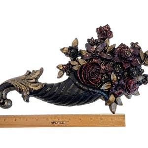 Puede incluir: Una cornucopia negra rebosante de flores burdeos, rojas y doradas. La cornucopia est&aacute; hecha de un material texturizado y tiene un acabado dorado. Las flores est&aacute;n dispuestas en un estilo realista y tienen un toque vintage.