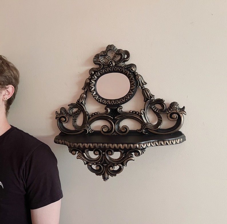 May include: A black and gold ornate wall shelf with a round mirror in the center. The shelf has a decorative design with scrolls and flourishes.