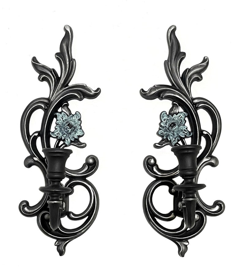 May include: A pair of black metal wall sconces with ornate floral designs and blue floral accents. Each sconce has a single candle holder.
