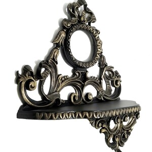 May include: A black and gold ornate wall shelf with a decorative scrollwork design. The shelf has a small, round opening in the center.