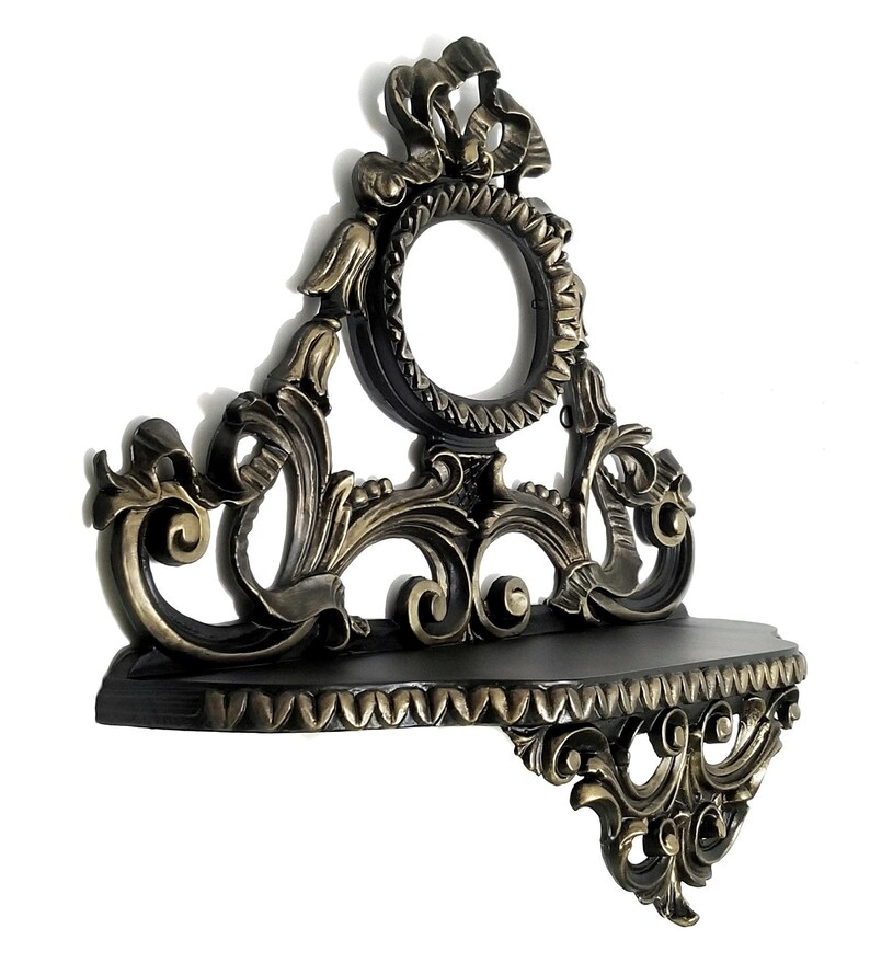 May include: A black and gold ornate wall shelf with a decorative scrollwork design. The shelf has a small, round opening in the center.