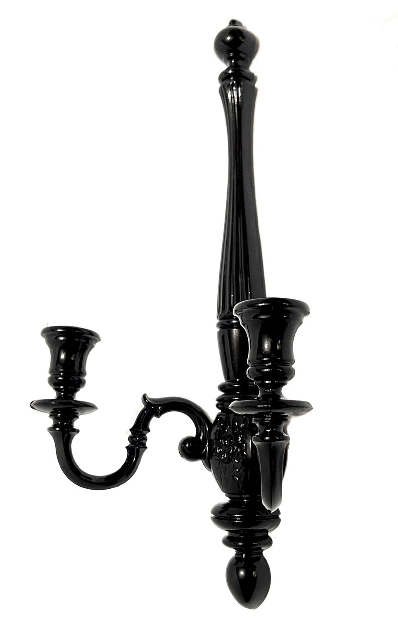 Op de afbeelding: Zwarte metalen wandkandelaar met twee armen en een decoratief ontwerp. De kandelaar heeft een hoge, slanke basis en twee gebogen armen die elk een kaarshouder dragen.