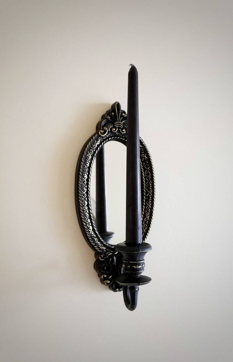 May include: Black candle holder with an oval mirror and ornate gold detailing. The candle holder is mounted on a white wall.