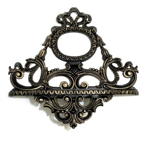 May include: A decorative wall shelf with an ornate, gold-painted, and black-painted design. The shelf has a scalloped edge and a central oval opening.