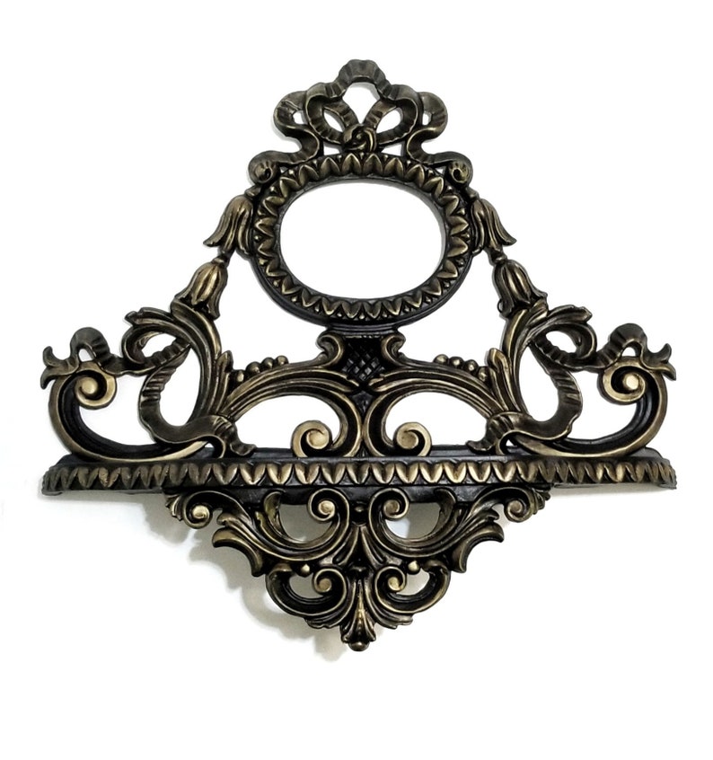 May include: A decorative wall shelf with an ornate, gold-painted, and black-painted design. The shelf has a scalloped edge and a central oval opening.