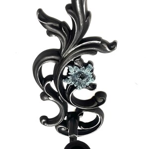 May include: A black metal candle holder with a decorative floral design and a blue glass flower accent. The holder has a black rubber base.