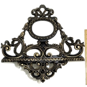 May include: A gold-toned metal wall shelf with ornate scrollwork and a decorative oval frame. The shelf has a black finish and a scalloped edge.