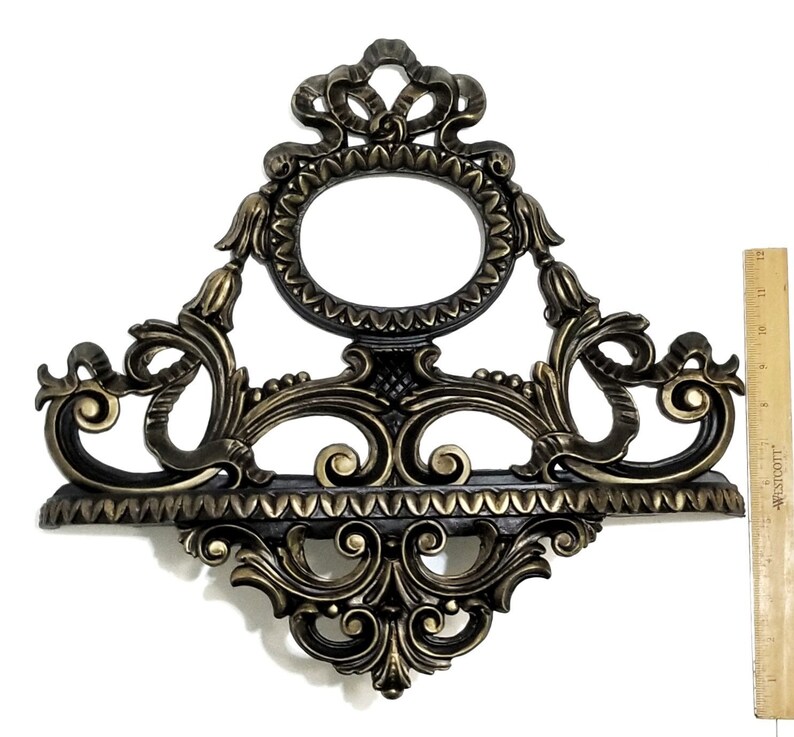 May include: A gold-toned metal wall shelf with ornate scrollwork and a decorative oval frame. The shelf has a black finish and a scalloped edge.