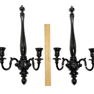 Op de afbeelding: Een paar zwarte metalen wandkandelaars met elk drie kaarsarmen. De kandelaars zijn versierd en hebben een decoratief ontwerp.