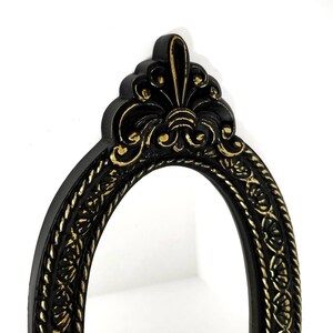 May include: Black oval mirror with a gold ornate frame. The frame has a decorative design with a fleur-de-lis at the top.