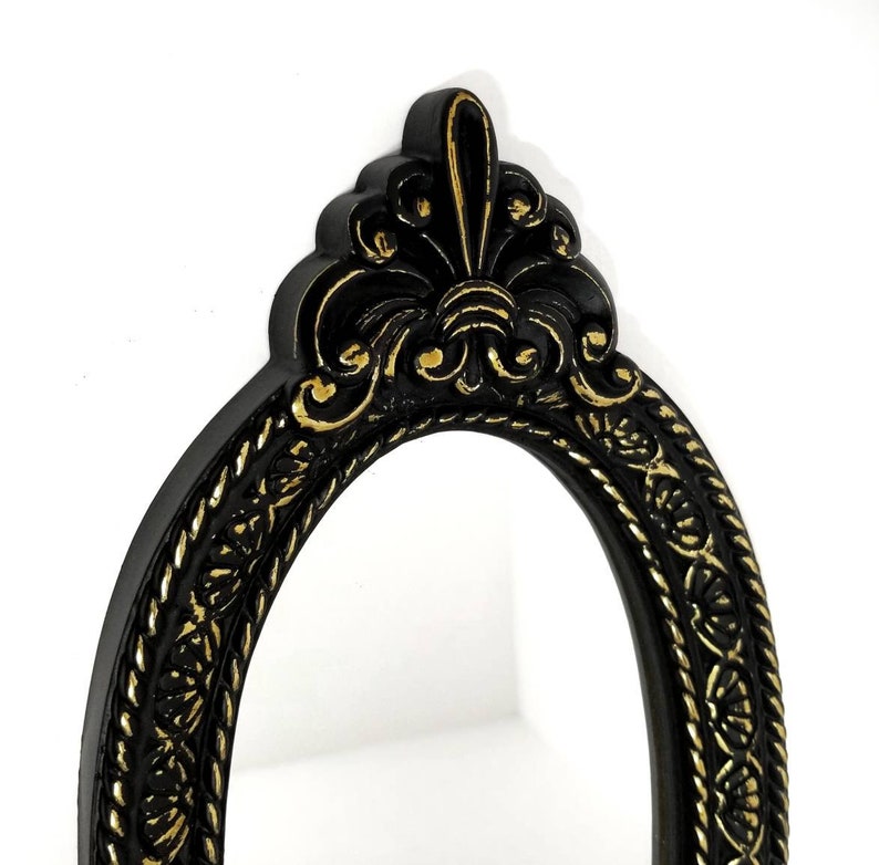 May include: Black oval mirror with a gold ornate frame. The frame has a decorative design with a fleur-de-lis at the top.