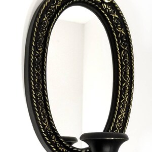 May include: Black oval mirror with a gold ornate frame and a single candle holder on the side. The mirror has a simple design and the candle holder is ornate.