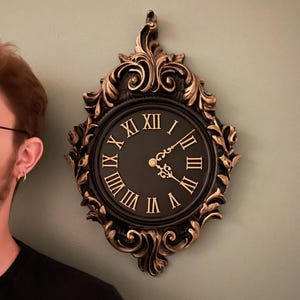 May include: A black and gold ornate clock with Roman numeral markings on the face. The clock has a decorative, scrolled frame.