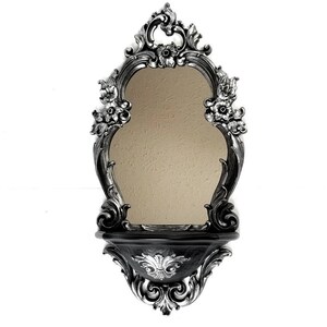 May include: A black and silver ornate wall mirror with a shelf. The mirror has a scalloped oval shape and is surrounded by intricate floral designs. The shelf is also decorated with floral designs.
