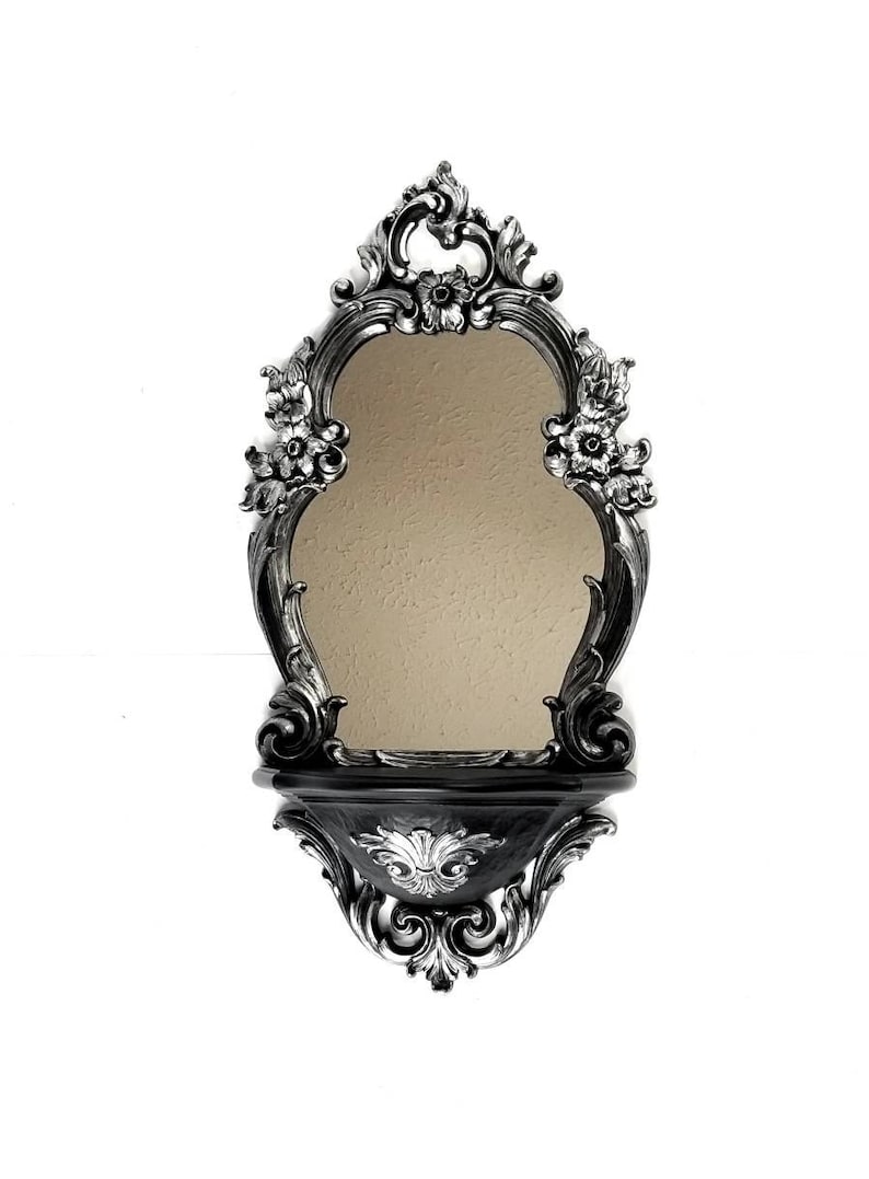 May include: A black and silver ornate wall mirror with a shelf. The mirror has a scalloped oval shape and is surrounded by intricate floral designs. The shelf is also decorated with floral designs.