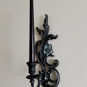 May include: Black metal wall sconce with ornate scrollwork and a single candle holder. The sconce features a decorative floral design with a blue and white flower.