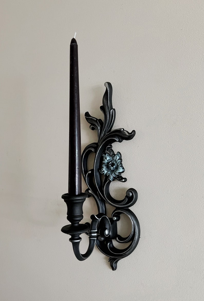 May include: Black metal wall sconce with ornate scrollwork and a single candle holder. The sconce features a decorative floral design with a blue and white flower.
