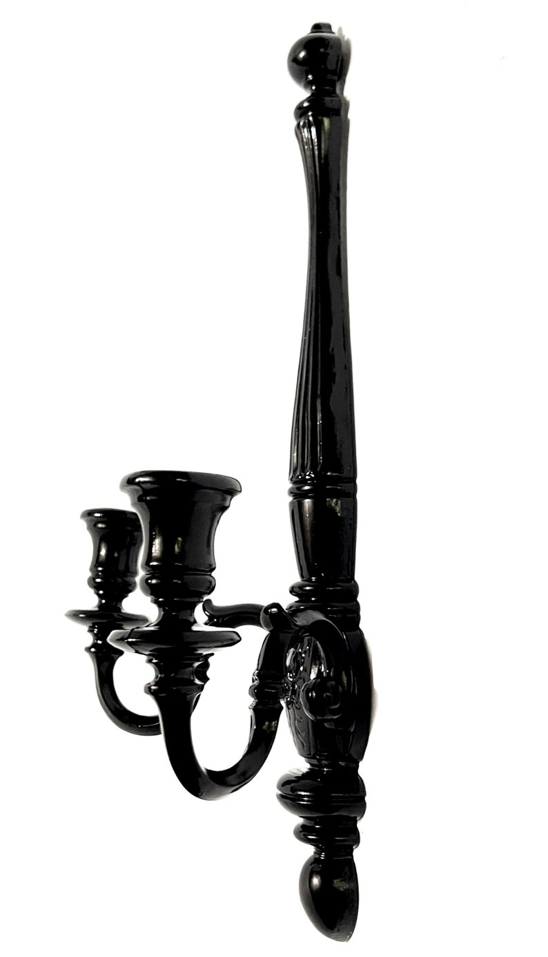 Op de afbeelding: Zwarte metalen wandkandelaar met drie kaarsarmen. De kandelaar heeft een decoratief ontwerp en een zwarte afwerking.