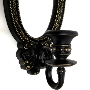 May include: Black wall-mounted candle holder with an oval mirror and ornate gold accents. The candle holder has a decorative design with a scroll-like shape and a ring for hanging.