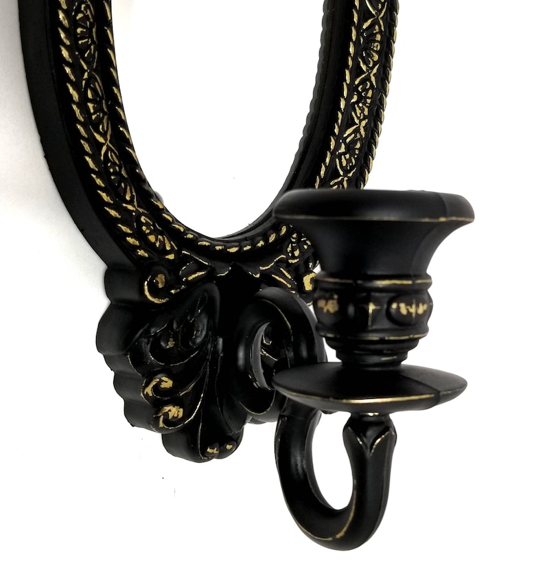 May include: Black wall-mounted candle holder with an oval mirror and ornate gold accents. The candle holder has a decorative design with a scroll-like shape and a ring for hanging.