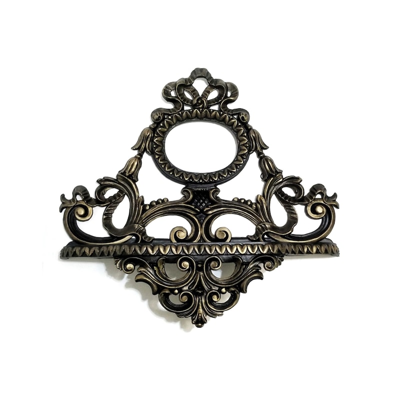 May include: A gold and black ornate wall shelf with a decorative, round, framed mirror in the center. The shelf has a scalloped edge and intricate scrollwork.