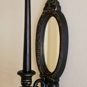 May include: Black wall-mounted candle holder with an oval mirror. The candle holder has gold accents and a decorative design. The mirror has a simple, plain surface.