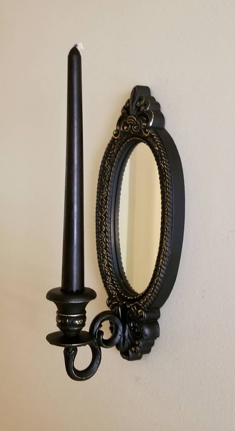 May include: Black wall-mounted candle holder with an oval mirror. The candle holder has gold accents and a decorative design. The mirror has a simple, plain surface.