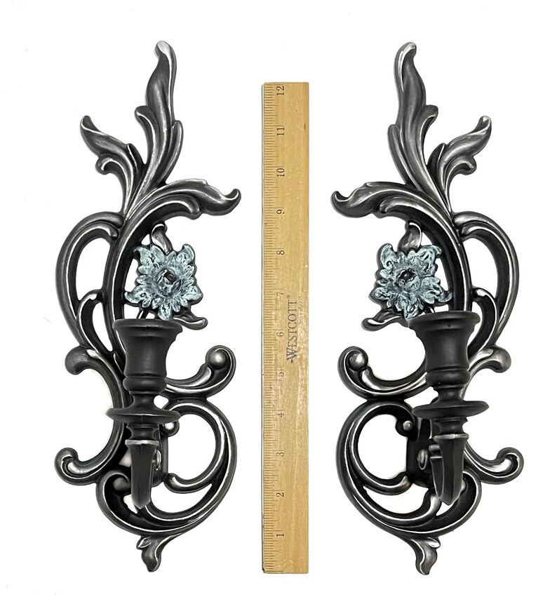 May include: A pair of silver metal wall sconces with a floral design and a blue glass flower accent. Each sconce has a candle holder.