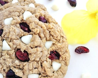 May include: Close-up of a chewy oatmeal cookie with white chocolate chips and dried cranberries. The cookie is on a white surface with a yellow background.