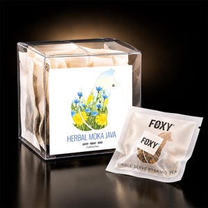 Puede incluir: Una caja de plástico transparente con bolsitas de té y una etiqueta con la silueta de un zorro rellena de flores azules y amarillas. La etiqueta dice HERBAL MOKA JAVA, EARTHY ROBUST SWEET, Caffeine Free. Una bolsita de té está al frente.