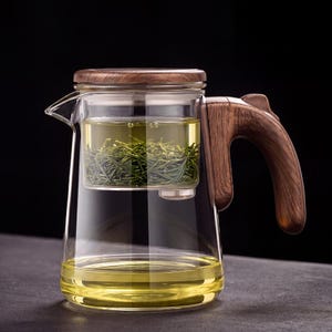 Peut inclure: Une théière en verre transparent avec une poignée et un couvercle en bois. La théière est dotée d'un infuseur intégré rempli de feuilles de thé vert. Le thé à l'intérieur est de couleur jaune clair, et la poignée et le couvercle sont en bois brun foncé.