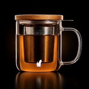 May include: A clear glass tea mug with a handle, featuring a removable stainless steel mesh infuser inside filled with amber liquid. The mug is topped with a circular wooden lid and displays a small white fox silhouette logo on the front surface.