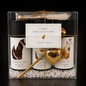 Może przedstawiać: Przezroczyste pudełko zawierające trzy cylindryczne puszki na herbatę z etykietami Chocolate Chai, Vanilla Chai i Indian Chai, każda z ilustracją lisa. Zestaw zawiera złoty zaparzacz do herbaty w kształcie serca, mieszadełko z cukrem i biały wypełniacz.