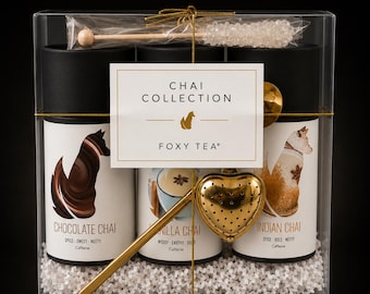 May include: A clear box containing three cylindrical tea canisters labeled Chocolate Chai, Vanilla Chai, and Indian Chai, each featuring a fox illustration. Includes a gold heart-shaped tea infuser, a rock candy stirrer, and white packing filler.