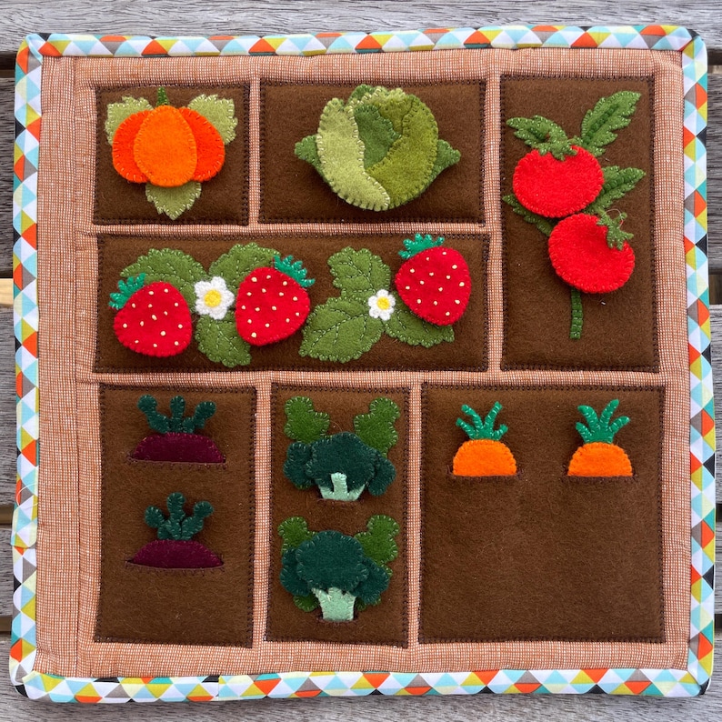 May include: A felt garden play mat with various vegetables and fruits, including a pumpkin, cabbage, tomatoes, strawberries, carrots, and broccoli. The mat is brown with a light brown border and a colorful patterned trim.