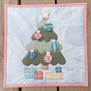 Peut inclure: Une décoration murale en forme de sapin de Noël en feutre avec une étoile au sommet et des ornements dans les tons bleus, roses et verts. L'arbre est entouré de cadeaux emballés dans du papier bleu, rose et jaune.